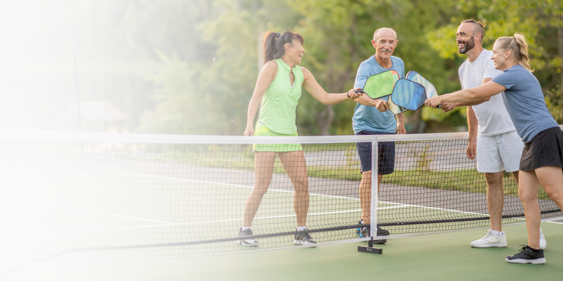 white gradient over a photo of a group of four adults who are tapping racquets to show sportsmanship after a Pickleball game together. They are each dressed comfortably in athletic wear and are smiling as they enjoy their time in the fresh air.